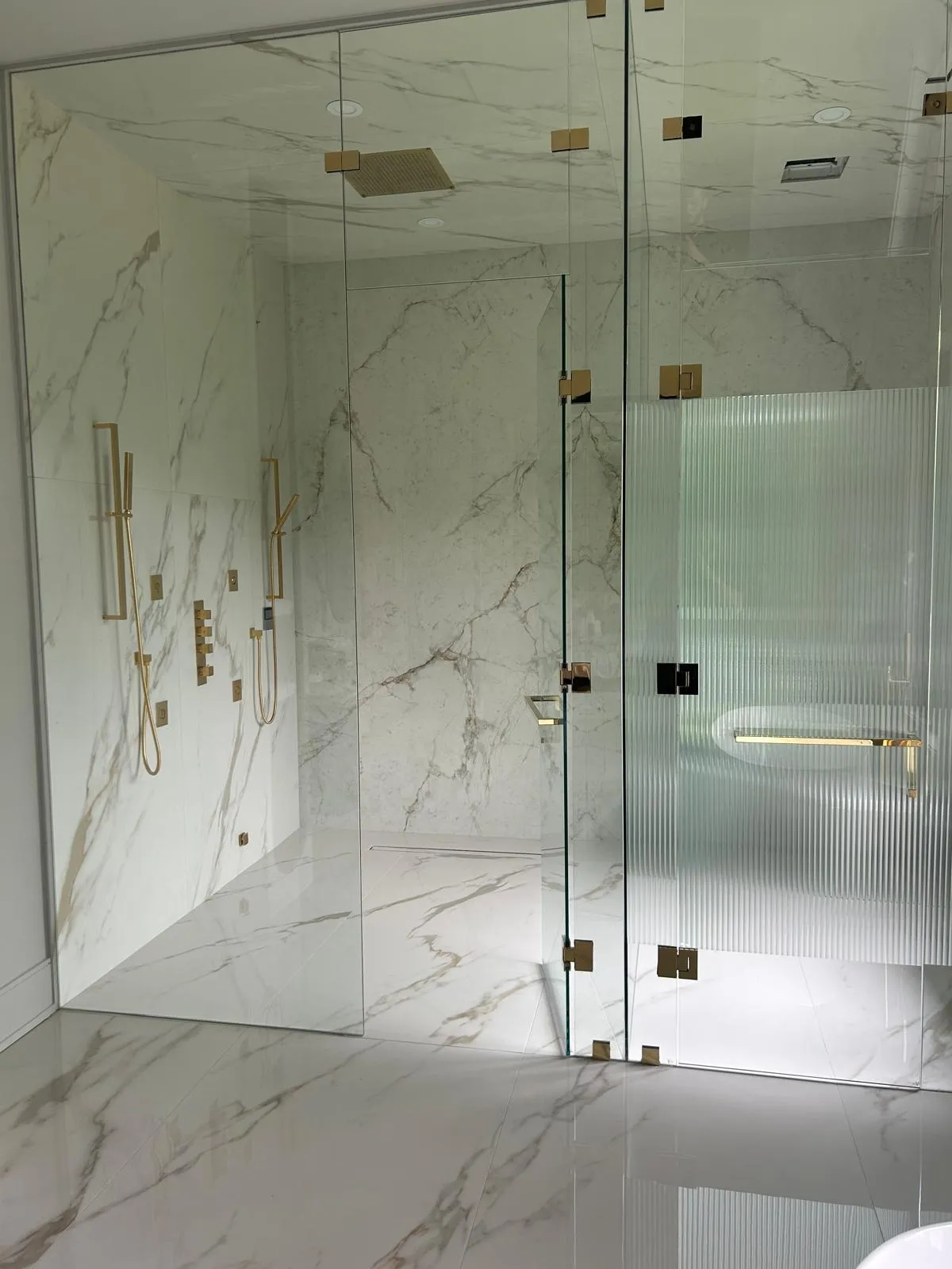 Curbless double shower in Whitby with book-matched Calacatta-effect porcelain, brass fixtures, a reeded glass panel and a linear drain
