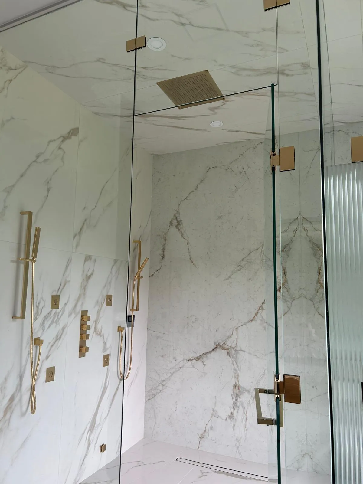 Book-matched Calacatta-effect porcelain running across the shower walls and ceiling, with brass body jets and a ceiling rain head, Whitby